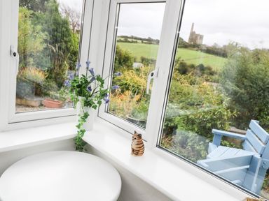 A corner window with a potted plant and small cat figurine overlooking a garden with a blue chair and a distant castle at Alice Cottage in Wheal Kitty near St Agnes