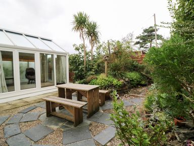 An outdoor patio with wooden benches and table next to a glass conservatory surrounded by plants and trees at Alice Cottage in Wheal Kitty near St Agnes