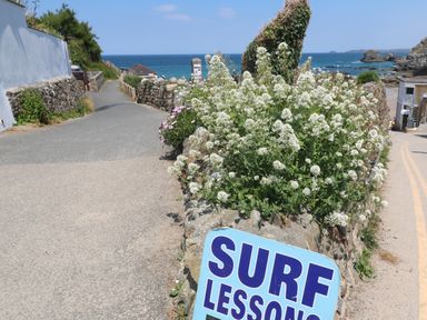 A coastal path with flowers and a surf lessons sign near the beach with rocks and ocean in the background at Alice Cottage in Wheal Kitty near St Agnes