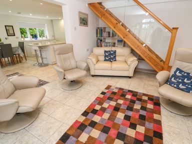 A living room with beige armchairs and sofa with patterned cushions a colorful checkered rug and a bookshelf under a wooden staircase at Alice Cottage in Wheal Kitty near St Agnes