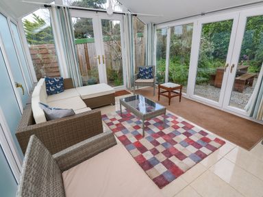 A sunroom with a wicker sectional sofa a glass coffee table a checkered rug and doors opening to a garden at Alice Cottage in Wheal Kitty near St Agnes