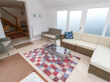 A sunroom with wicker seating including a sofa and chairs a glass coffee table and a multicolored checkered rug at Alice Cottage Wheal Kitty near St Agnes