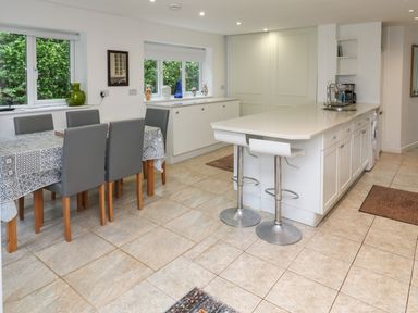 A kitchen with a dining table and chairs a countertop with bar stools and cabinets with windows showing greenery at Alice Cottage in Wheal Kitty near St Agnes