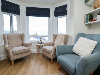 A sitting area with two beige armchairs and a blue chair with a white cushion by three windows with dark blinds and a small round table with wine glasses and a bottle at Mariners Watch in Whitby