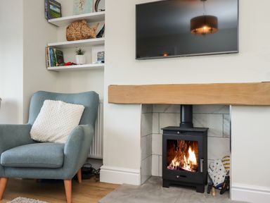 A blue armchair with a white pillow next to a wood burning stove in a fireplace with a wooden mantel and a wall-mounted TV above at Mariners Watch in Whitby