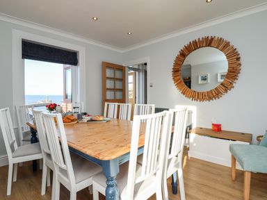 A dining room with a wooden table surrounded by white chairs next to a window with sea view and a round wooden framed mirror on the wall at Mariners Watch in Whitby