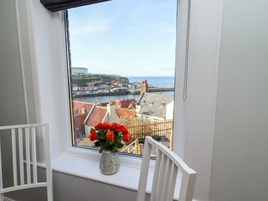 A vase with orange flowers on a windowsill with white chairs and a view of rooftops and sea at Mariners Watch in Whitby