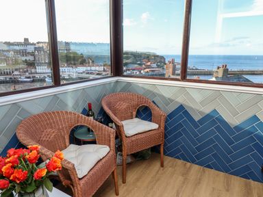 A corner with two wicker chairs with cushions a small round table with a wine bottle and glass and a vase with orange and red flowers by windows overlooking a coastal town at Mariners Watch in Whitby