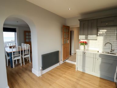 A kitchen with grey cabinets a sink and flowers on the counter next to a dining area with a wooden table and white chairs at Mariner's Watch in Whitby