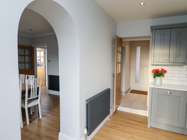 A kitchen with grey cabinets and a vase of flowers on the counter next to a hallway with a wooden door and an archway leading to a dining area with white chairs and a wooden table at Mariners Watch in Whitby