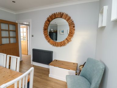 A dining area with a wooden table and white chairs a round wooden framed mirror a gray radiator a wooden chest and a blue chair at Mariners Watch in Whitby