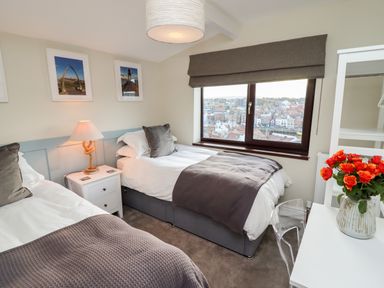 A bedroom with two single beds a nightstand with a lamp a window with a view of buildings and a desk with a vase of orange flowers at Mariners Watch in Whitby