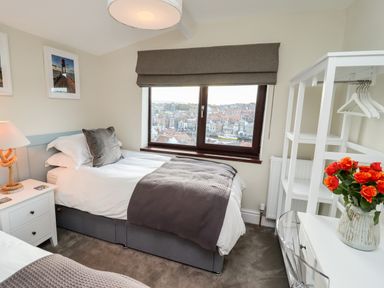 A bedroom with two single beds a window with a view a nightstand a lamp a white shelving unit and a vase with red flowers at Mariners Watch in Whitby
