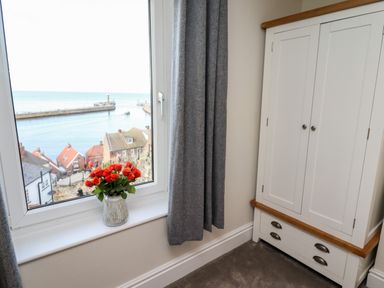 A window with grey curtains showing a view of rooftops and the sea with a vase of red flowers on the windowsill next to a white wardrobe at Mariners Watch in Whitby