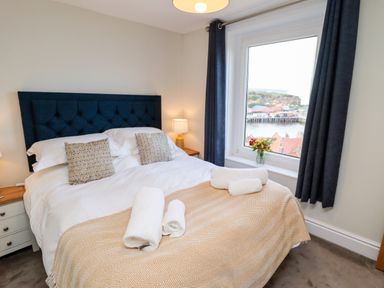 A bedroom with a double bed with a blue headboard two patterned pillows bedside tables with lamps a window with dark curtains and a view of a harbor at Mariners Watch in Whitby