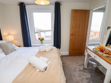 A bedroom with a bed with towels on it a window with curtains a door and a desk with breakfast items at Mariners Watch in Whitby