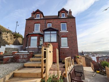 A brick house with dormer windows and wooden steps leading to the entrance overlooking a town and water at Mariners Watch in Whitby