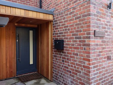 Entrance with a dark door set in wooden cladding beside a brick wall with a mounted mailbox at Mariners Watch in Whitby
