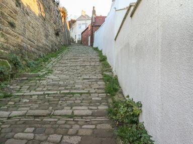 A steep cobblestone street with a stone wall on the left and a white wall on the right leading to buildings at Mariner's Watch in Whitby