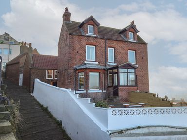 A two-story brick house with white window frames and a front porch beside a sloped cobblestone pathway at Mariner's Watch in Whitby