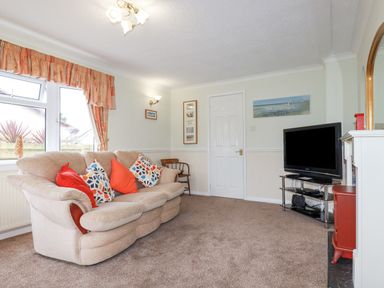A living room with a beige sofa with colorful cushions a television on a stand and a window with curtains at Summer Breeze in Tregolds near St Merryn