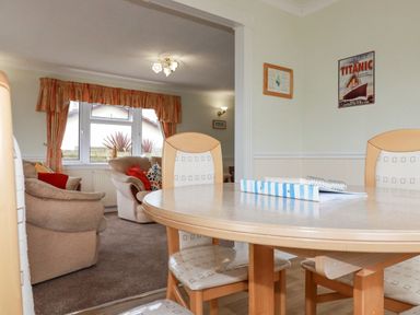 A dining area with a round table and chairs next to a living room with beige armchairs and a window with curtains at Summer Breeze in Tregolds near St Merryn