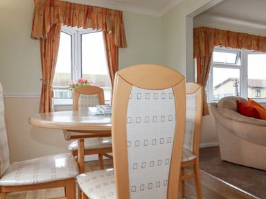 A dining area with a wooden dining table and padded chairs beside windows with curtains at Summer Breeze in Tregolds near St Merryn