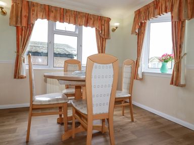 A dining area with wooden table and four chairs near two windows with orange curtains and a vase of flowers at Summer Breeze in Tregolds near St Merryn