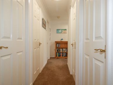 A hallway with closed white doors on both sides a wooden bookshelf at the end and paintings on the walls at Summer Breeze Tregolds near St Merryn