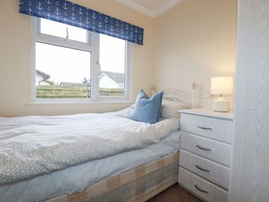 A bedroom with a single bed a blue pillow a bedside table with a lamp and a window with blue anchor patterned blinds at Summer Breeze in Tregolds near St Merryn