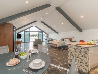 An open plan living space with dining table set with food a kitchen island with stone base a bed seating area and large windows overlooking a lake at Boathouse on the Lake in Bowness-On-Windermere
