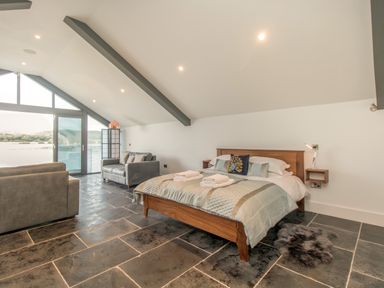 A bedroom with a wooden bed, two gray sofas, stone tile floor, and large windows showing a lake view at Boathouse on the Lake in Bowness-On-Windermere