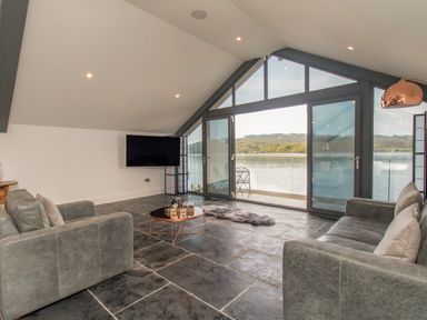 A living room with gray sofas a round coffee table a wall-mounted TV and large windows overlooking a lake at Boathouse on the Lake in Bowness-On-Windermere