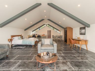 A spacious loft room with a bed, armchair, coffee table, dining table, desk, and kitchen area at Boathouse on the Lake in Bowness-On-Windermere