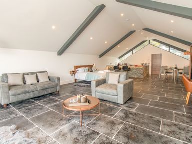 An open-plan room with a gray sofa and armchair around a round coffee table a bed near the back and a kitchen with bar stools and a dining area at Boathouse on the Lake in Bowness-On-Windermere