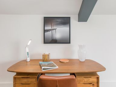 A wooden desk with a lamp tissue box book coaster and vase with chair and framed photo on wall at Boathouse on the Lake in Bowness-On-Windermere