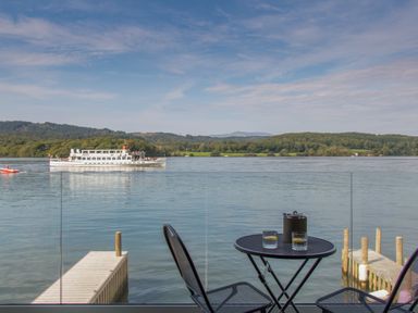 A lake view with a boat on the water and two chairs and a table with drinks on a balcony at Boathouse on the Lake in Bowness-On-Windermere