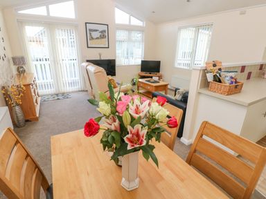A dining table with a vase of flowers in a living room with sofas televisions and a cabinet at The Hideaway - Heritage Park in Stepaside near Kilgetty