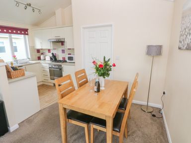 A dining area with a wooden table and four chairs next to a kitchen with white cabinets and appliances at The Hideaway - Heritage Park in Stepaside near Kilgetty
