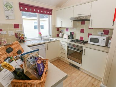A kitchen with white cabinets a gas stove an oven a microwave a wine rack a sink under a window and a wicker basket with food items on the counter at The Hideaway - Heritage Park Seaside near Kilgetty