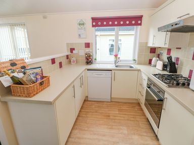 A kitchen with white cabinets and countertops a window with a red valance above the sink and a wicker basket with snacks and a bottle on the counter at The Hideaway - Heritage Park in Stepaside near Kilgetty