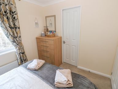 A bedroom with a bed, towels on the bed, a wooden dresser with a mirror and a basket, and a closed white door at The Hideaway - Heritage Park, Stepaside near Kilgetty