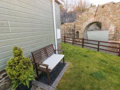 A wooden bench with a white cushion next to a potted plant by a house wall with a stone arch and fence in the background at The Hideaway - Heritage Park in Stepaside near Kilgetty