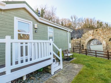 An exterior view of a green cabin with white railings and a bench on a lawn near stone arches at The Hideaway - Heritage Park in Stepaside near Kilgetty