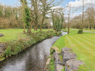 A narrow stream flows through a grassy area with trees and a rocky edge at The Hideaway - Heritage Park in Stepaside near Kilgetty