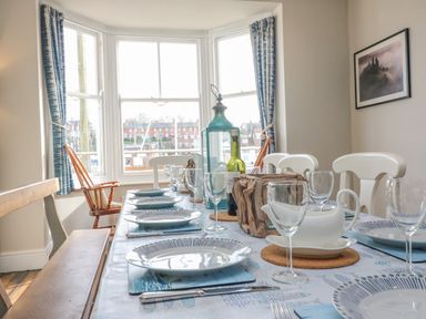 A dining table set with plates glasses and cutlery near bay windows with blue curtains at Seasides in Weymouth