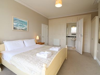 A bedroom with a double bed, bedside table with lamp, wall painting, and an open door showing a bathroom at Seasides in Weymouth