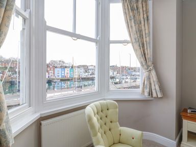 A corner of a room with a light green armchair in front of a large window overlooking a harbor with boats and colorful buildings at Seasides in Weymouth