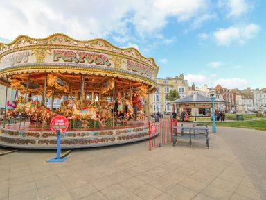 An outdoor carousel with people riding horses near a kiosk and benches in a town square at Seasides in Weymouth