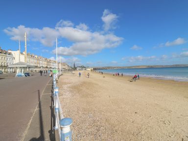 A beach with people walking along the sandy shore and a promenade with buildings by the side at Seasides in Weymouth